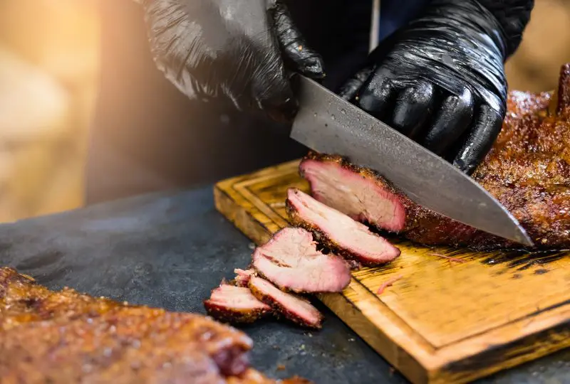 Gloved hands slicing smoked brisket on a wooden cutting board.